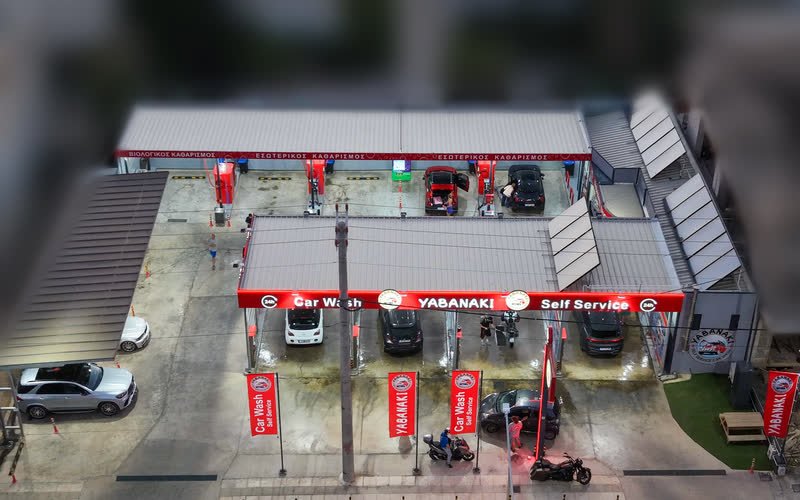 Λήψη από ψηλά του Yabanaki Self Service Car Wash: κόκκινο φωτιζόμενο στέγαστρο με την επιγραφή Car Wash Yabanaki Self Service, τέσσερις θέσεις πλύσης με αυτοκίνητα και κόκκινες σημαίες με το σήμα